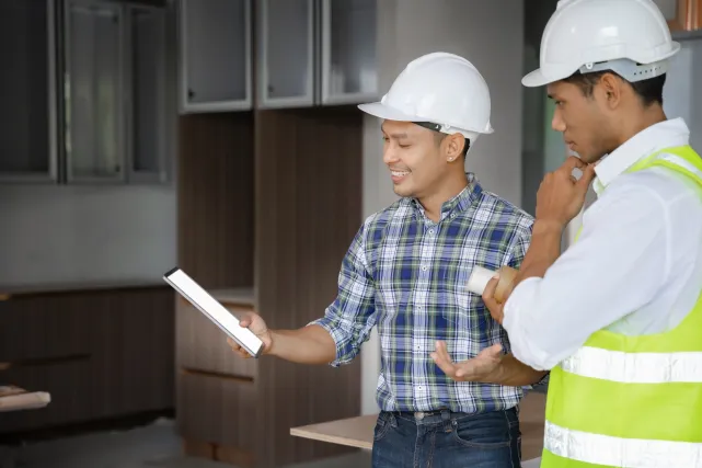 Two workers in hard hats review plans inside a building.