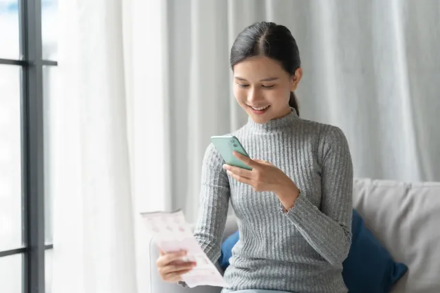 A woman uses a smartphone while holding a paper document.