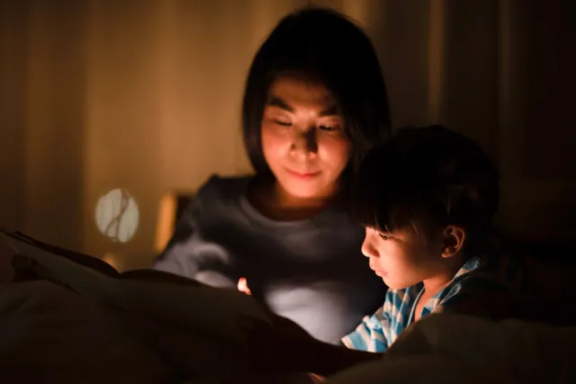 A woman and child read together by a light in a dark room.