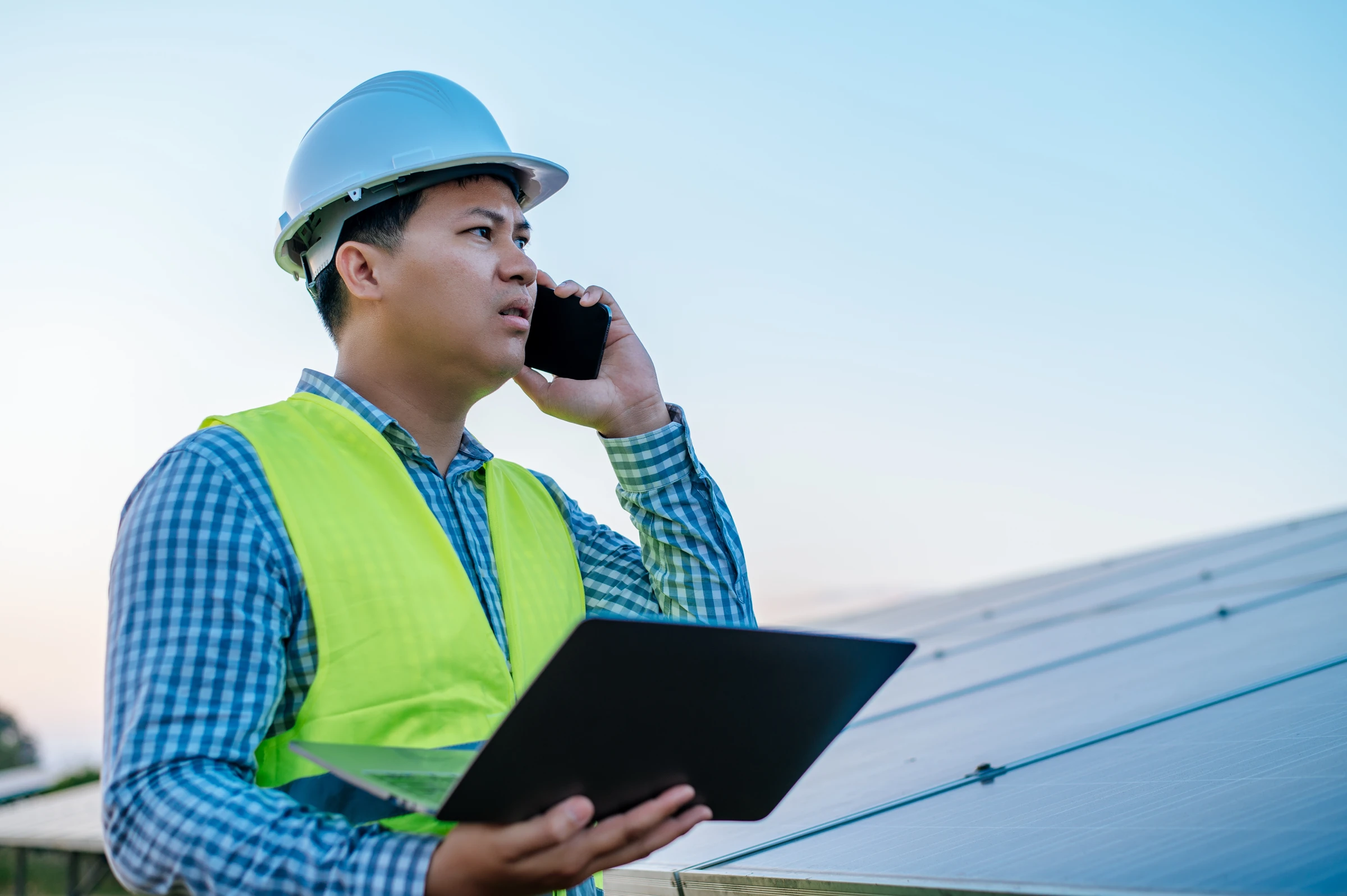 A worker in a hard hat holds a laptop and speaks on a phone beside solar panels.