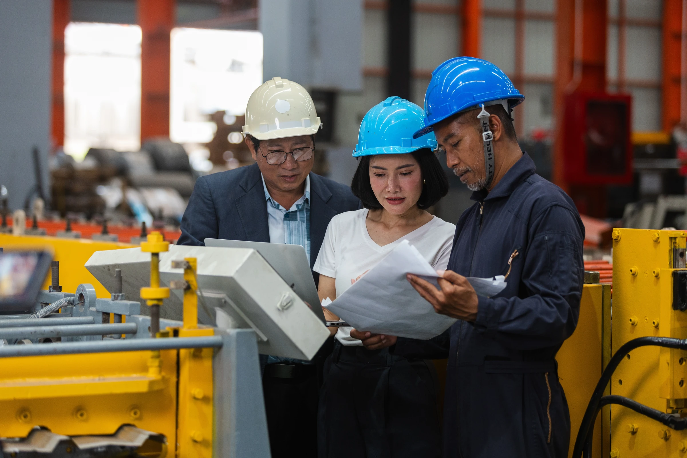 Three people in hard hats review a laptop and documents in a factory.