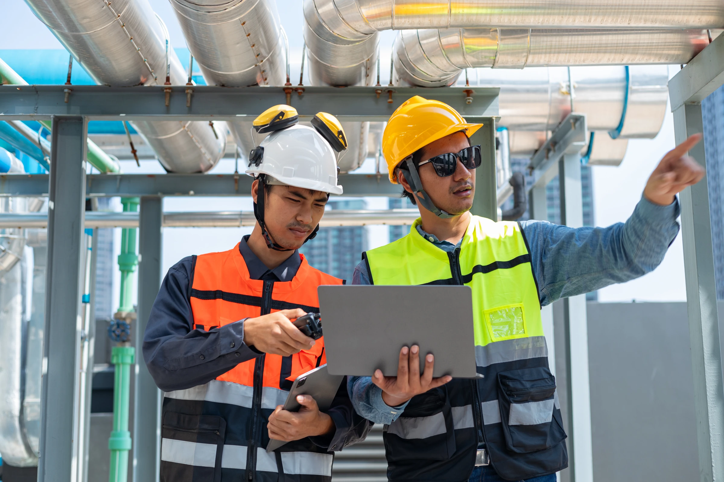 Two workers in hard hats review a laptop beside industrial equipment.
