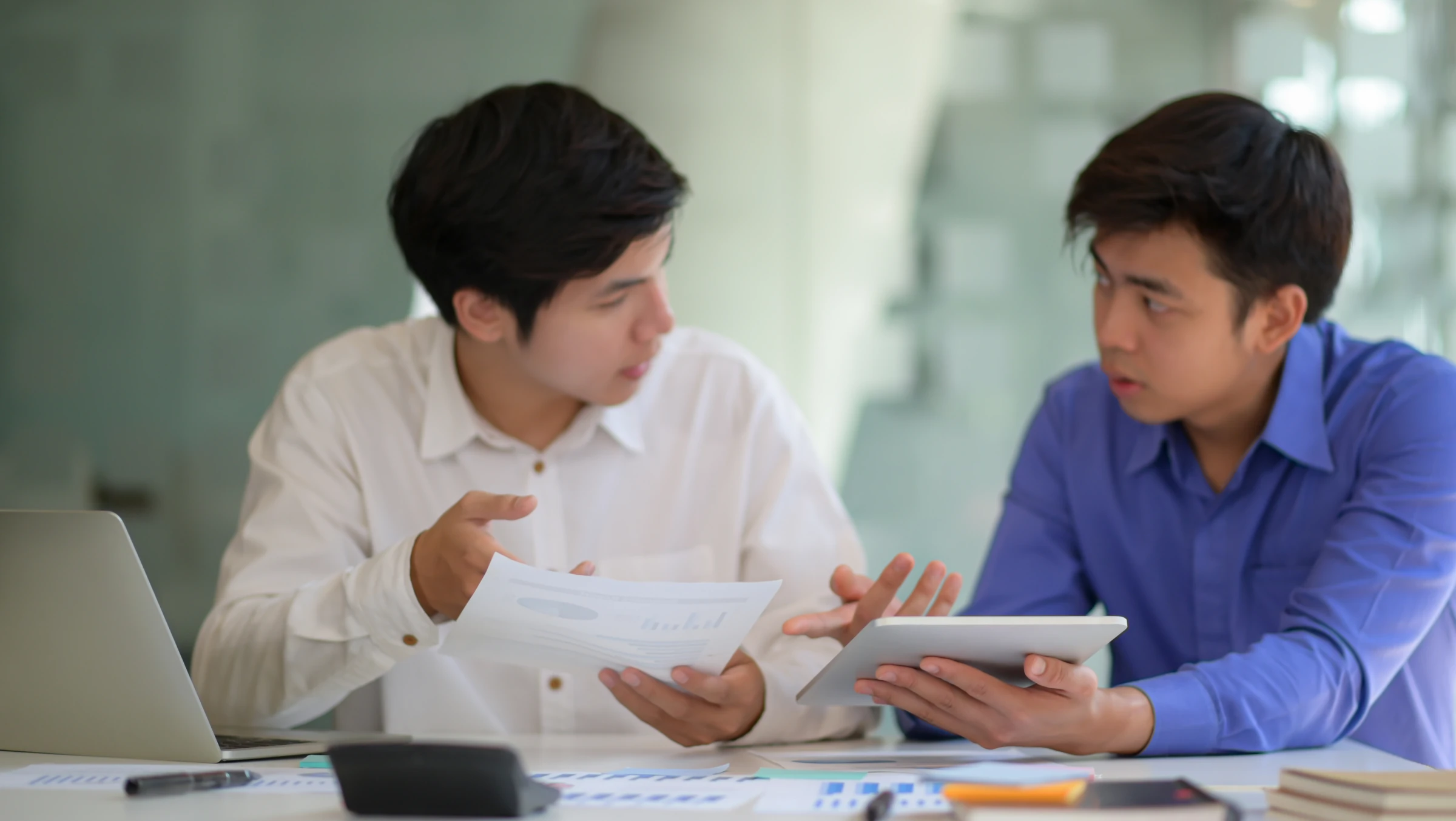 Two men discuss documents and a tablet at a desk.