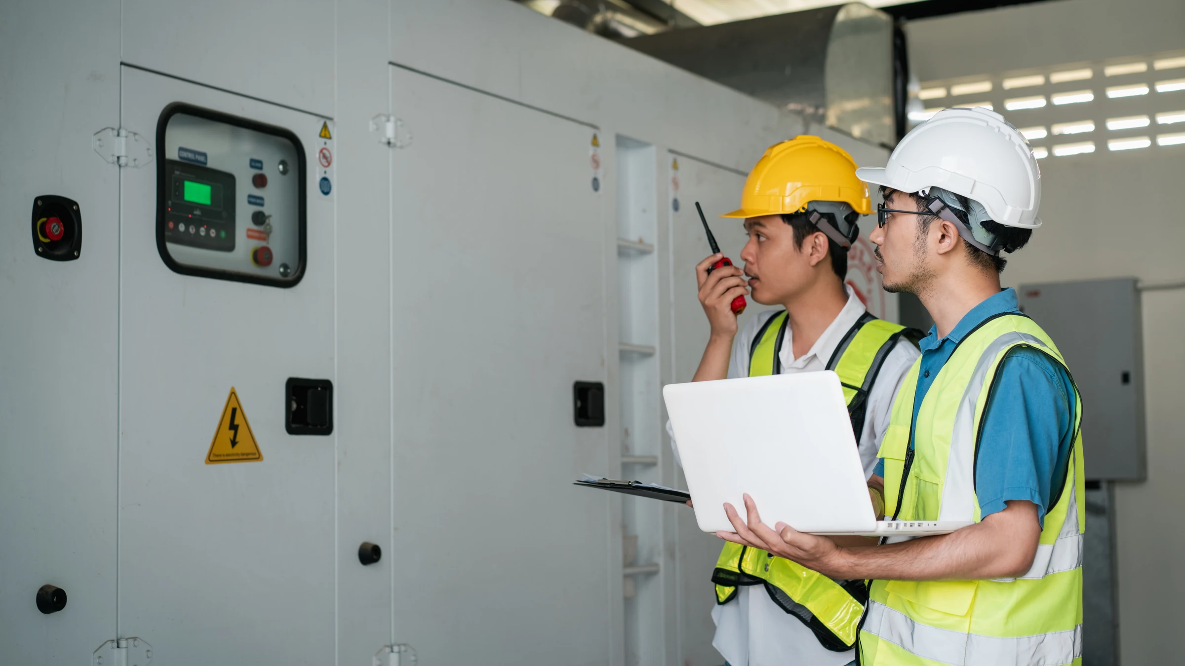 Two workers in hard hats inspect electrical control equipment.