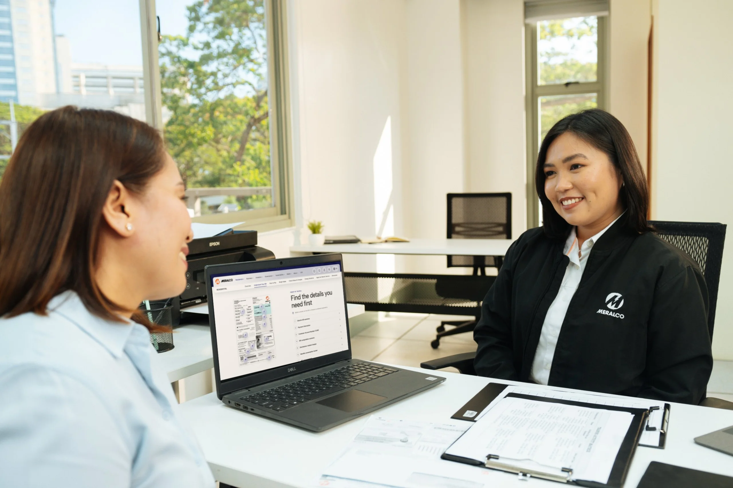 A Meralco representative assists a customer beside a laptop displaying the Meralco website.