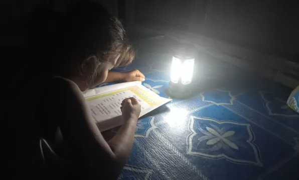 A student writes in a workbook beside a glowing solar lamp in a dark room.