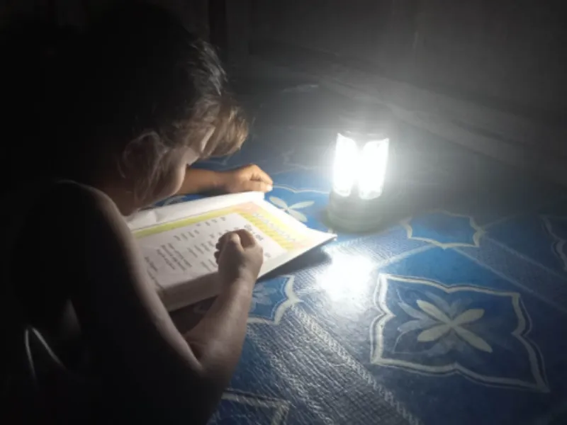 A student writes in a workbook beside a glowing solar lamp in a dark room.