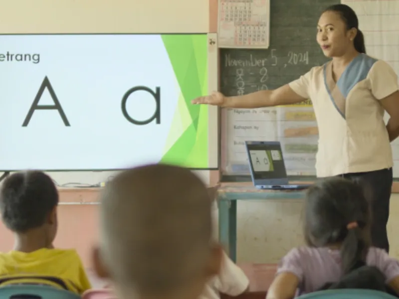 A teacher points to letters on a classroom screen while students watch.