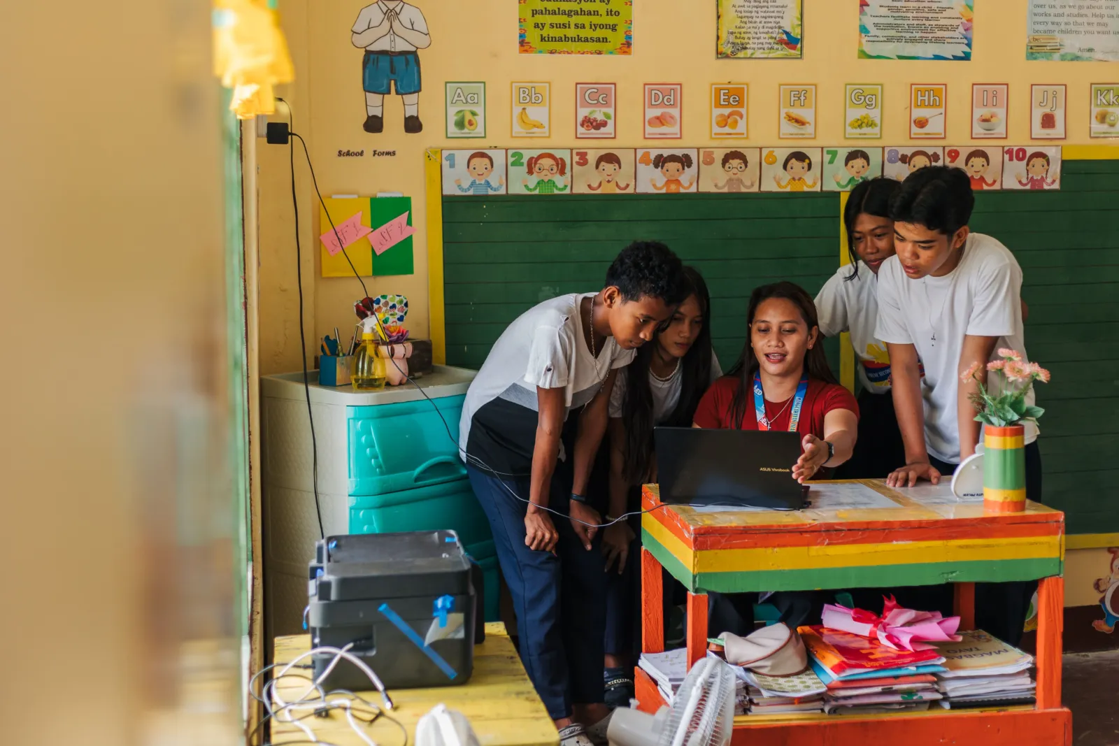 Students gather around a teacher using a laptop in a classroom.