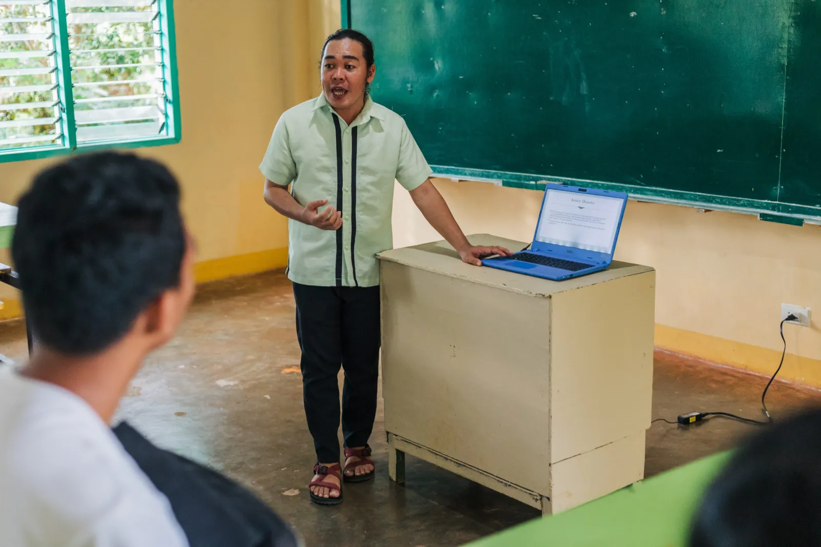Jonel Elnar teaches beside a laptop and chalkboard while students listen.