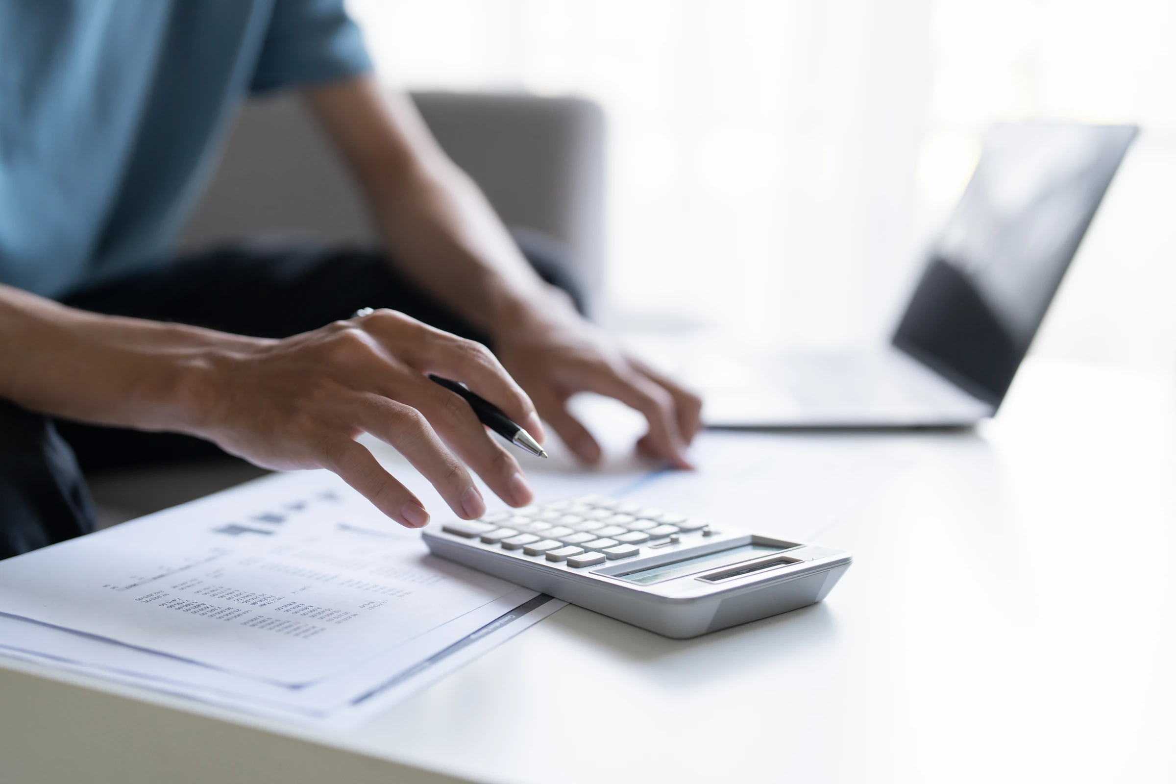 A person uses a calculator beside paperwork and a laptop.