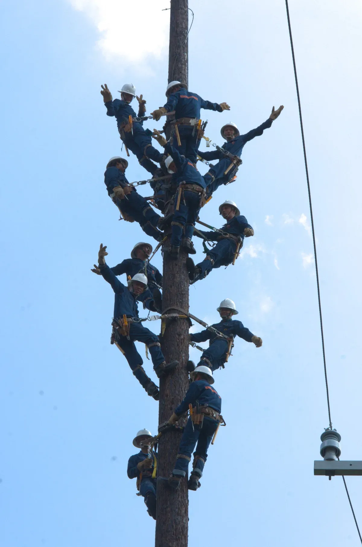Meralco linemen in white hard hats pose along a utility pole against a blue sky.